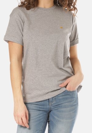 Kobieta w gładkim szarym T-shircie z krótkim rękawem i niebieskich dżinsach, z jedną ręką w kieszeni i widocznymi kręconymi włosami.