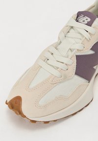 Baskets New Balance beige et blanches avec logo latéral violet, lacets blancs et semelle en caoutchouc texturée marron sur fond blanc.