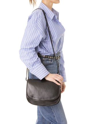 Zwarte leren schoudertas met een gebogen flap, gladde textuur en verstelbare band; casual gedragen met een blauw gestreept shirt en jeans.