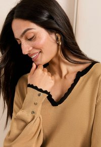 Pull en beige avec un col et des poignets ondulés noirs, comportant trois boutons décoratifs en or sur la manche gauche. Texture en tricot douce.