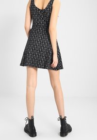 Černé šaty bez rukávů s bílými geometrickými vzory, rozšířenou sukní a kulatým výstřihem, doplněné černými kotníkovými botami.