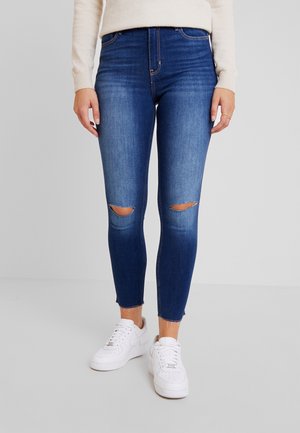 Femme portant un jean skinny bleu déchiré et des baskets blanches, debout avec les mains détendues le long du corps sur un fond clair uni.