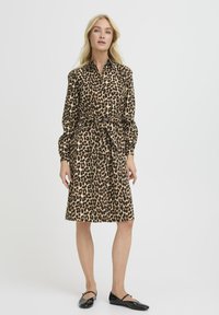 Srajčna obleka s leopardjim potiskom in dolgimi rokavi, z oblečenim pasom, ki vsebuje ovratnik in gumbe. Material se zdi mehak z gladko teksturo.