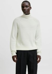 Suéter blanco de cuello alto con canalé y mangas raglán, combinado con pantalones negros a rayas. Textura suave y corte relajado.
