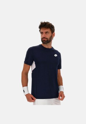 Mann trägt ein marineblaues kurzärmeliges Sportshirt mit Logo, weiße Handgelenksbänder und weiße Shorts, steht vor einem schlichten hellen Hintergrund.