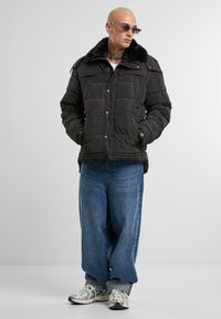Svart boblejakke med myk, kunstig pelskrage, frontlommer og trykknap-lukking. Brukt sammen med baggy blå jeans og sølvfargede joggesko.