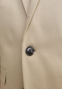 Μπεζ σακάκι με σκούρο κουμπί τεσσάρων τρυπών στερεωμένο κοντά στον γιακά και τσέπη με καπάκι στο ύψος της μέσης.