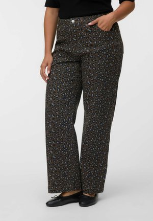 Femme portant un pantalon large taille haute à motif léopard marron et noir, et des ballerines noires, une main dans la poche.