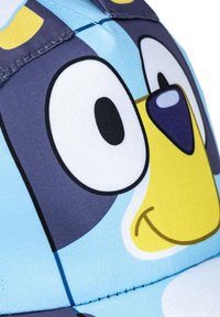 Niebieska czapka z wzorem postaci z kreskówki o dużych oczach i żółtym ryjku. Wykonana z teksturowanej tkaniny z widocznymi detalami szycia.