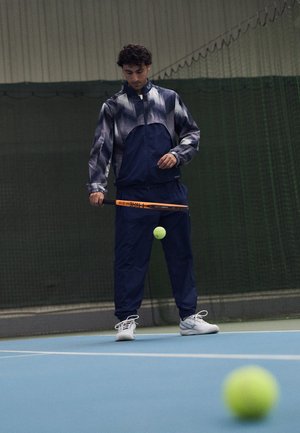 Granatowy dres sportowy z szarym geometricznym wzorem, połączony z białymi trampkami. Piłka tenisowa w powietrzu, a w ręku trzymany jest rakiet.