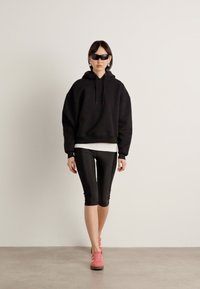 Crni oversized dukserica u kombinaciji s crnim tajicama i ružičastim tenisicama. Model nosi elegantne crne sunčane naočale i stoji protiv neutralne pozadine.