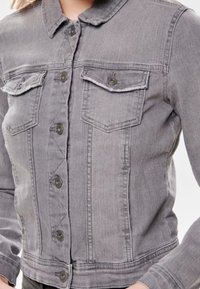 Chaqueta de denim gris con cuello tipo camisa, cierre frontal con botones, dos bolsillos en el pecho y una silueta ajustada con un ligero desgaste.
