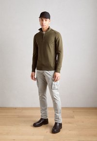 Sweater zippé vert olive, pantalon cargo gris avec poches latérales et bottes marron foncé. Le modèle porte une casquette noire, debout sur un sol en bois.