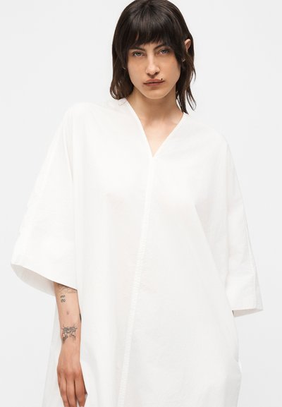 Άτομο με σκούρα μαλλιά φοράει άνετο λευκό φόρεμα με V λαιμόκοψη, δείχνοντας τατουάζ στα χέρια και σκουλαρίκι στα χείλη, πάνω σε απλό ανοιχτό φόντο.