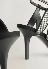 Sandales à talons hauts noires avec une finition brillante, dotées de brides perforées ornées de petits clous argentés et d'une plateforme noire texturée.