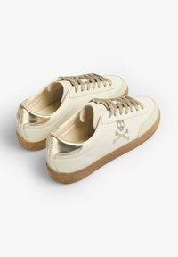 Weiße Ledersneaker mit goldenen Akzenten, ausgestattet mit einer strukturierten, braunen Sohle und goldenen Schnürsenkeln. Totenkopf- und Kreuzbones-Logo an der Seite.