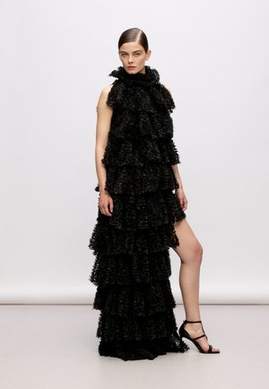 Black ruffled dress with a high neckline and layered design. The fabric has a textured appearance, paired with strappy black heeled sandals.