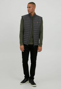 Chaleco gris acolchado con cuello alto y ribetes negros, llevado sobre una camisa de manga larga verde, combinado con pantalones negros y zapatillas negras.