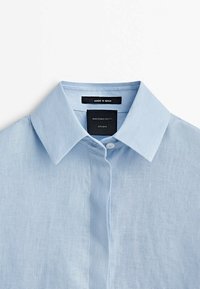 Chemise en lin bleu clair avec un col classique, une fermeture à boutons sur le devant et une étiquette d'entretien, présentant une texture subtile. Fabriquée en Espagne.