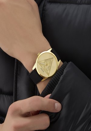 Orologio color oro con quadrante decorato e cinturino in silicone nero, indossato su una mano davanti a uno sfondo di giacca a vento nera.