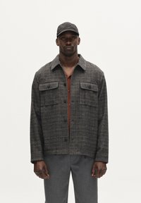 Hallide ruuduline jakk, valmistatud tekstuurilisest kangast, nelja esikotiga ja terava kraega, kihtide kaupa kantud roostes värvi särgi peal.