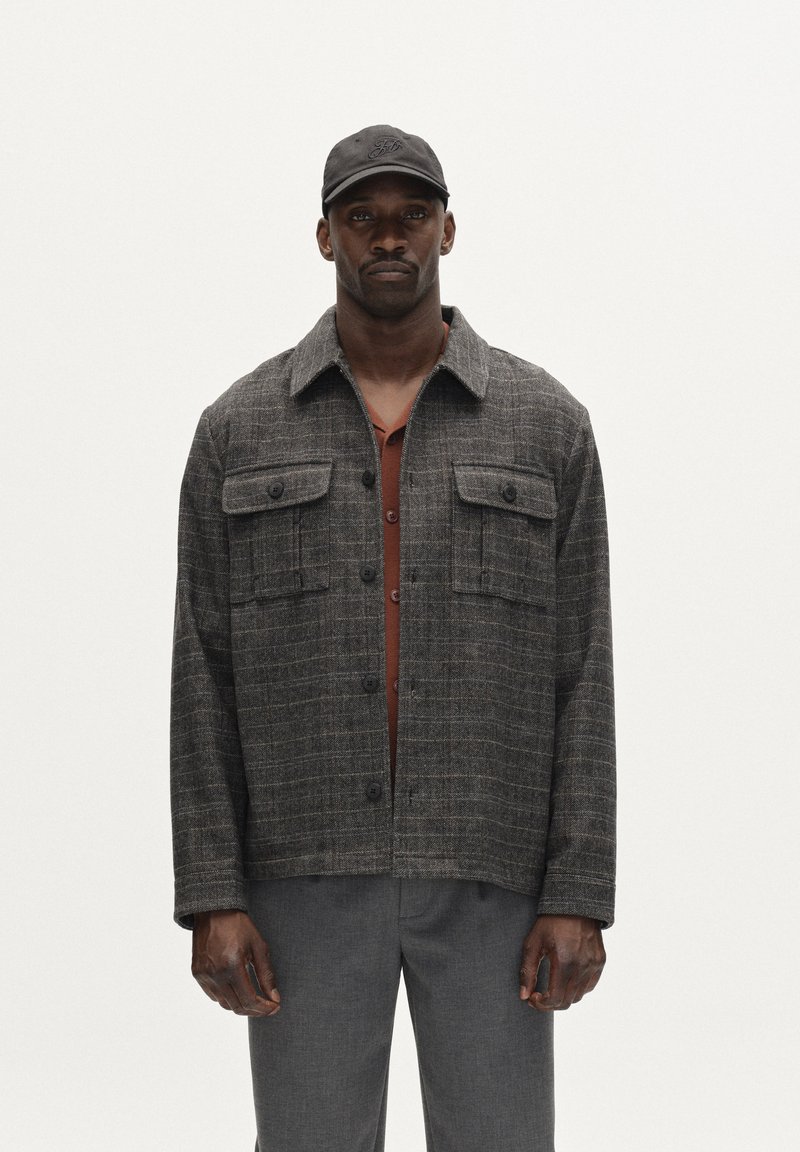 Hallide ruuduline jakk, valmistatud tekstuurilisest kangast, nelja esikotiga ja terava kraega, kihtide kaupa kantud roostes värvi särgi peal.