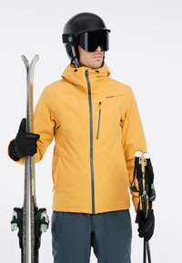 Geel skijas met een voorrits, verstelbare capuchon en zijzak. Zwarte handschoenen, ski's en stokken. Een zwarte helm en skibril maken de look compleet.