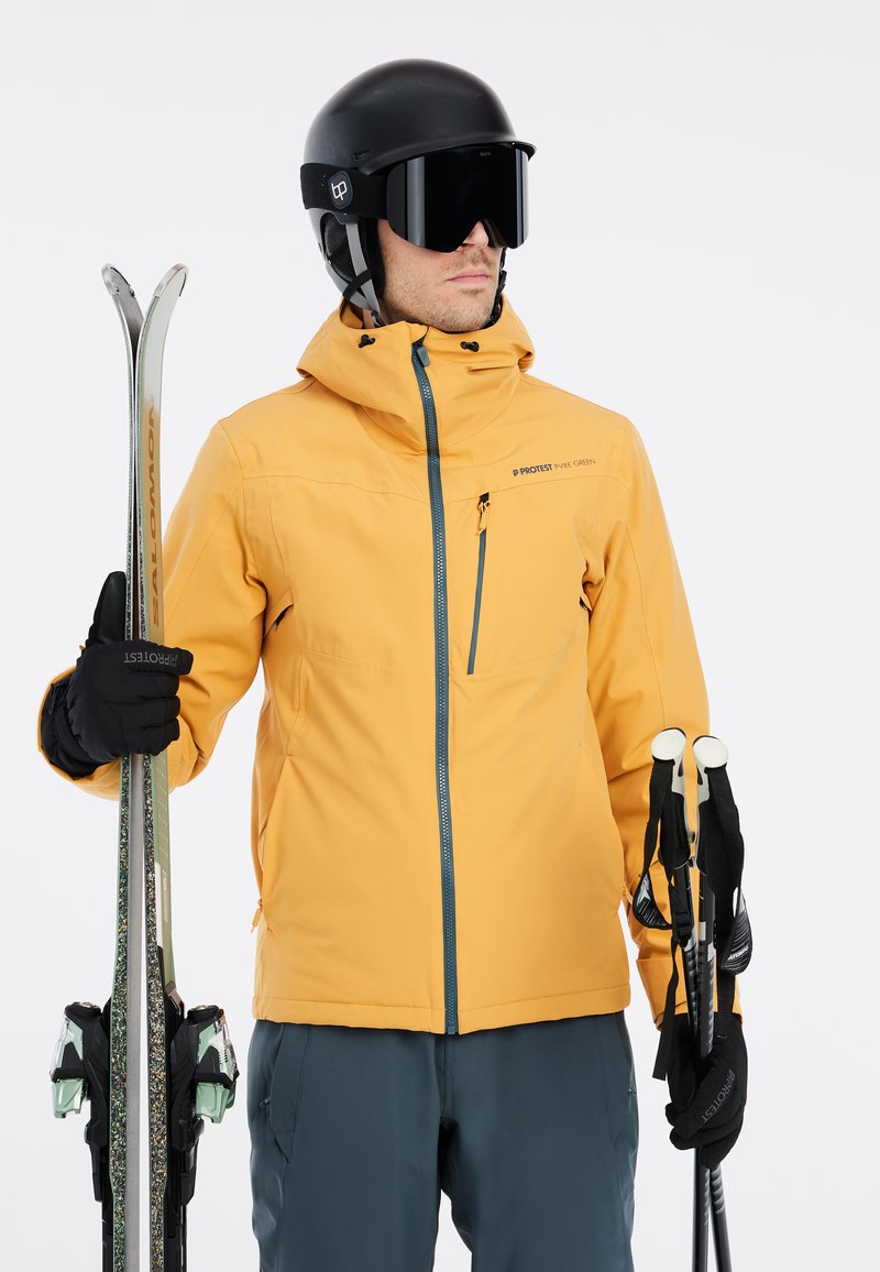 Geel skijas met een voorrits, verstelbare capuchon en zijzak. Zwarte handschoenen, ski's en stokken. Een zwarte helm en skibril maken de look compleet.