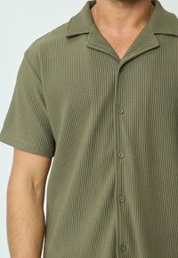 Chemise à manches courtes olive en tissu texturé à côtes, avec un col et une fermeture à boutons sur le devant.