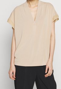 Beige kortärmad topp med V-ringning och avslappnad passform. Slät textur, minimal sömnad och en subtil fickdetalj på framsidan.