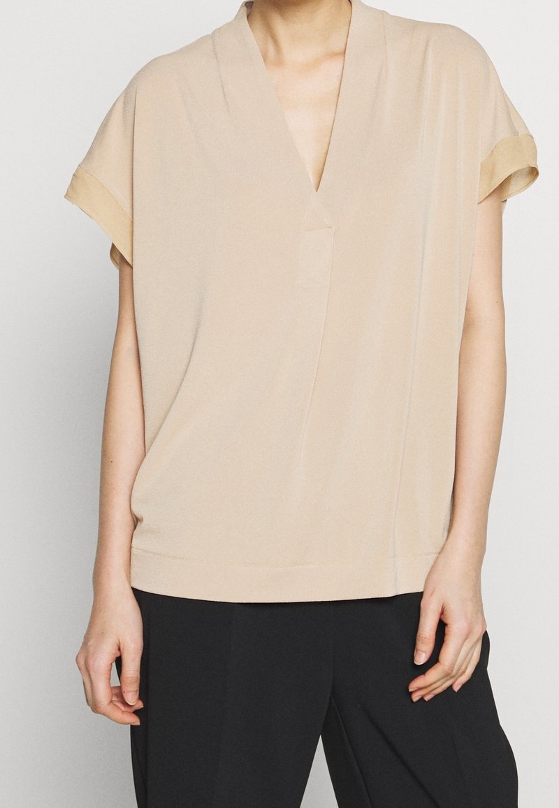 Beige kortärmad topp med V-ringning och avslappnad passform. Slät textur, minimal sömnad och en subtil fickdetalj på framsidan.