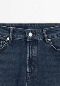 Jeans en denim bleu foncé avec une texture lisse, dotés d'une fermeture à bouton, de rivets en métal et de surpiqûres orange contrastantes le long des coutures.