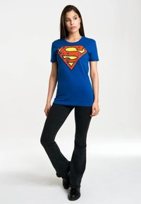 Blue cotton t-shirt featuring a large red and yellow Superman logo at the center. Paired with flared black jeans and black boots.