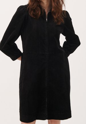 Femme portant une robe noire en velours côtelé mi-mollet avec des manches longues bouffantes et une fermeture éclair à l'avant, mains posées sur les hanches.