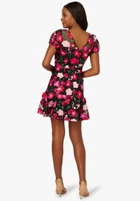 Květinové šaty s černým pozadím zdobené růžovými a červenými květy, krátkými nafouklými rukávy, výstřihem do V na zádech a volánkovým lemem. Materiál působí lehce.