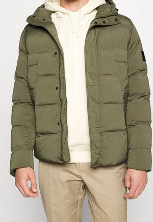 Mand iført olivengrøn dunjakke med hætte over beige hættetrøje og lysebrune bukser, stående mod en ensfarvet baggrund.