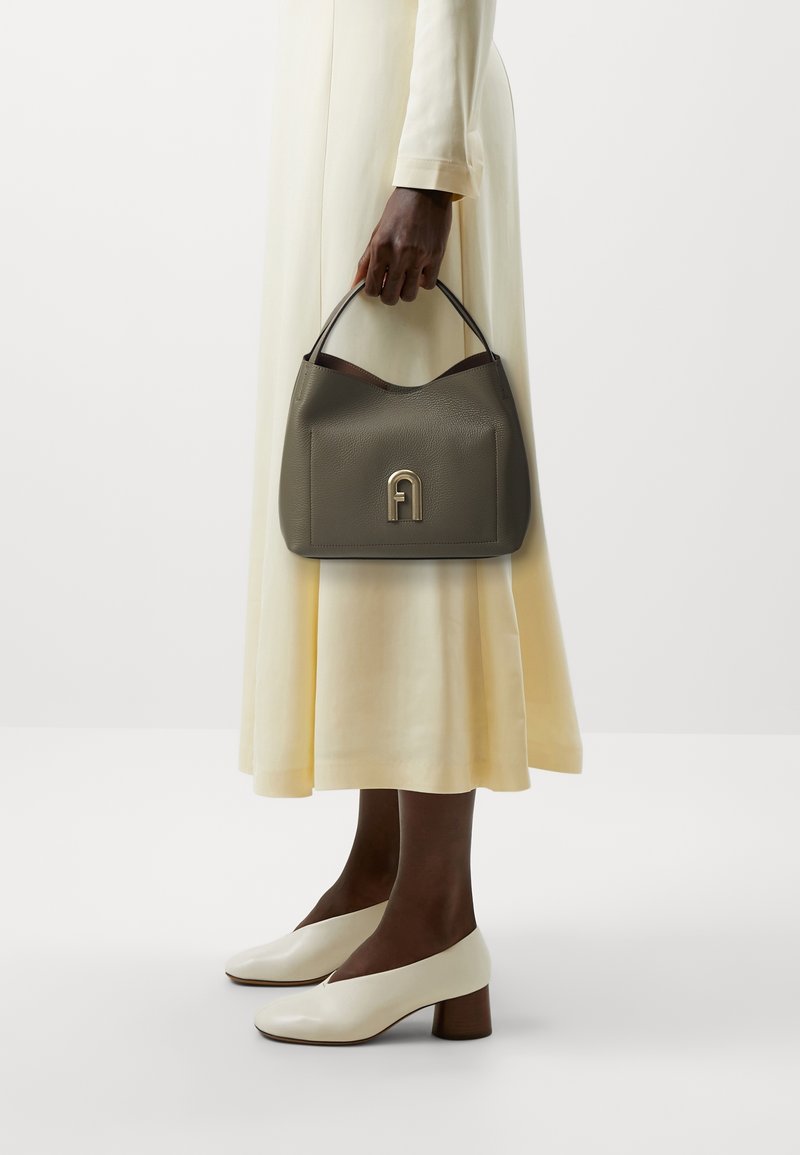 Personne portant une robe crème et des chaussures crème à talons bas tenant un sac à main gris avec un fermoir doré sur un fond blanc.