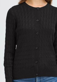Cardigan en tricot noir avec une texture en câble, un col rond et une fermeture à cinq boutons. Les manches longues et les poignets côtelés complètent le design.