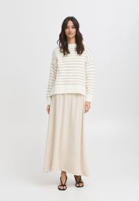 Pull rayé en crème et beige doux, assorti à une longue jupe beige fluide. Le pull présente une coupe décontractée et des poignets côtelés. Sandales noires.