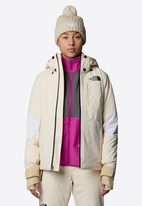 Giacca bianca sporca con accenti a blocchi di colore in grigio, rosa e bianco, con un alto colletto e zip frontale. Accompagnata da un berretto lavorato a maglia crema.