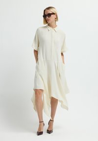 Vestido blanco de camisa de manga corta con delante abotonado, dobladillo asimétrico y bolsillos laterales. La modelo lleva gafas de sol oscuras y tacones marrones.