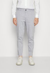 Man wearing light gray striped slim trousers, white dress shirt, and white lace-up shoes standing against plain background.