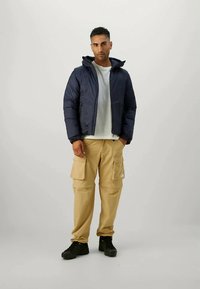 Navy puffer jacket with a hood, a front zipper, and elastic cuffs. Paired with beige cargo pants featuring pockets and black sneakers.