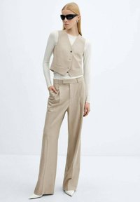 Beige taillierter Weste mit drei Knöpfen, kombiniert mit hochgeschnittenen, weiten Hosen. Das Model trägt ein Langarmoberteil und eine Sonnenbrille.