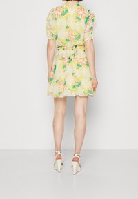 Květinové šaty ve žluté barvě s růžovými a zelenými akcenty. Mají nabírané rukávy, stažený pas a rozšířenou sukni, doplněné bílými podpatky.