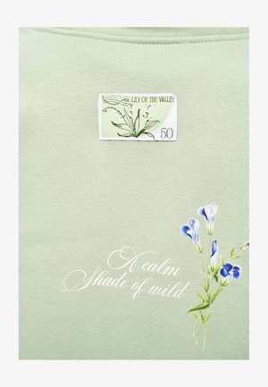 Jasnozielona tkanina z botaniczną naszywką oznaczoną "Konwalia 50", kursywnym napisem "Spokojny odcień dzikości" oraz małymi fioletowymi kwiatami.