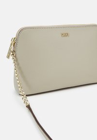 Beżowy portfel z syntetycznej skóry z złotym zamkiem, wytłoczonym logo i odpinanym złotym paskiem na ramię. Gładka faktura, minimalistyczny design.