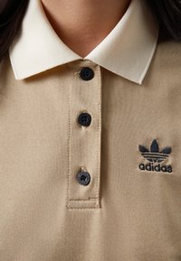 Béžové polo tričko s krémovým límečkem, třemi černými knoflíky a černým vyšitým logem Adidas na levé straně. Texturovaná látka.