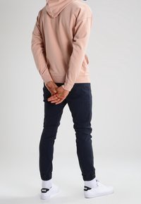 Felpa rosa chiaro con cappuccio e cordino, pantaloni jogger blu navy con polsini elastici e sneaker bianche con marchio nero sul tallone.