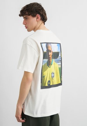 Ung mand med kort brunt hår iført hvid T-shirt med stort rektangulært billede af en fodboldspiller i gul brasiliansk trøje på ryggen.